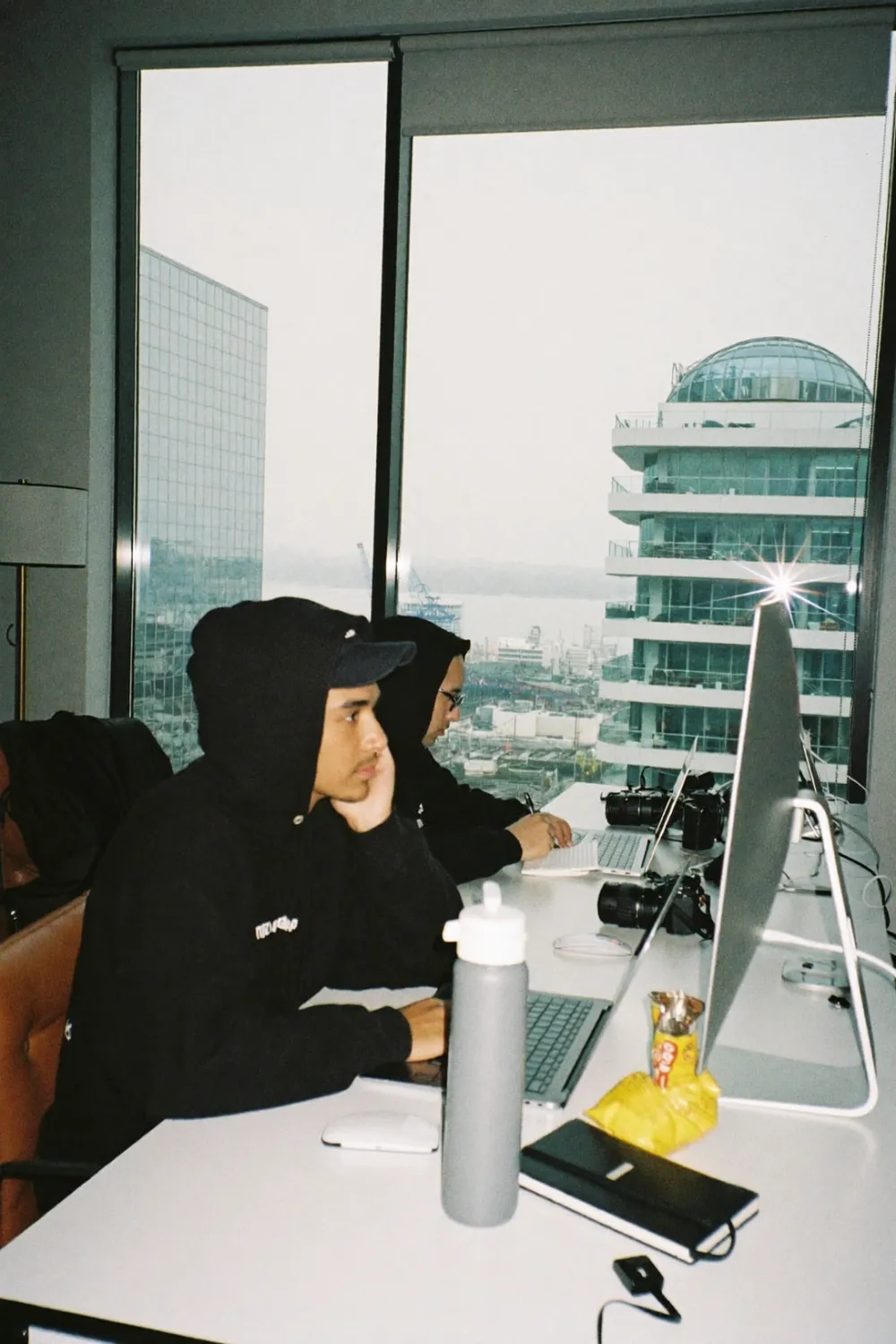 Two people in hoodies working quietly at laptops by a high-rise office window
