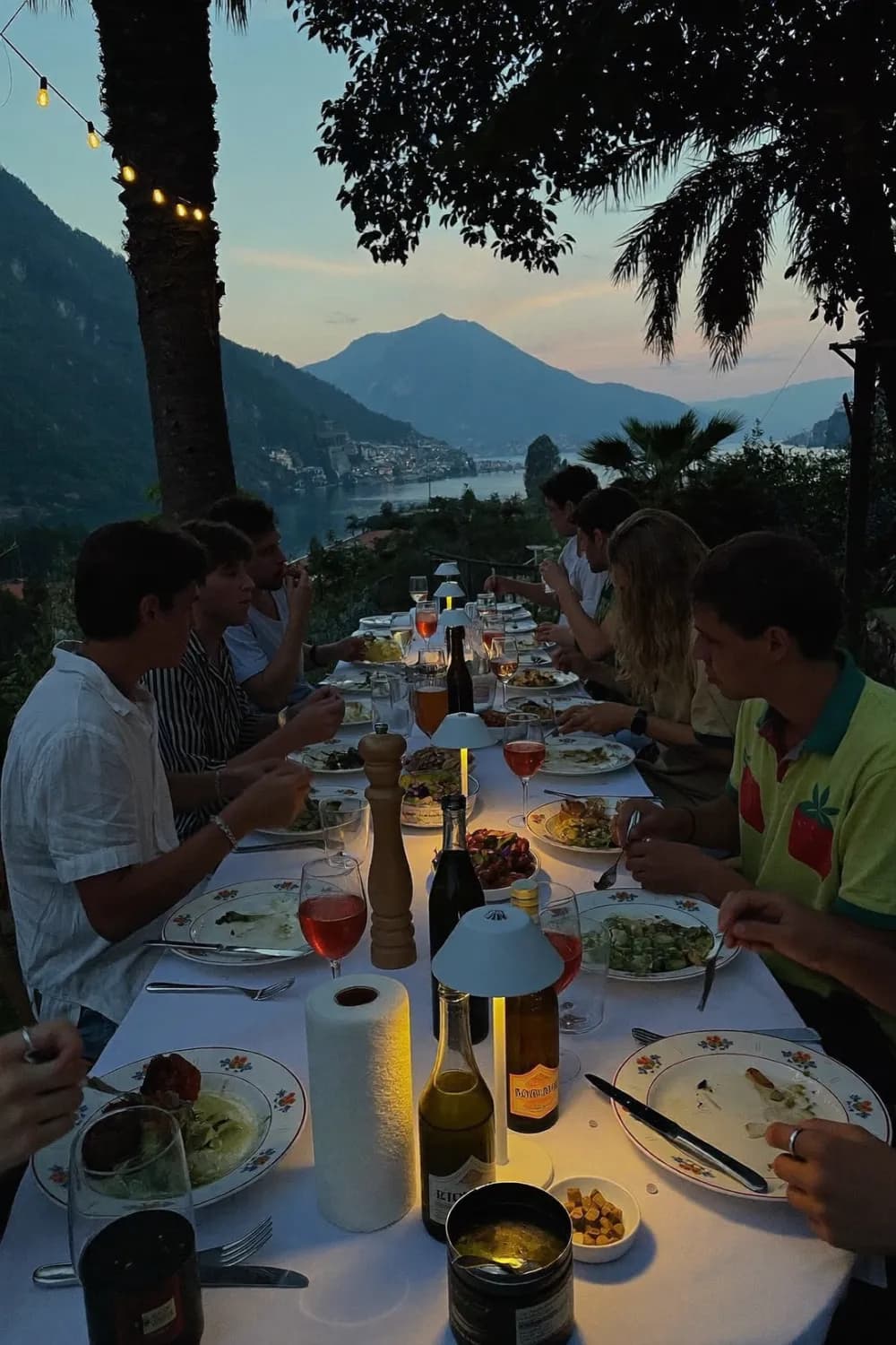Friends sharing a long outdoor dinner at dusk above a lake and mountains