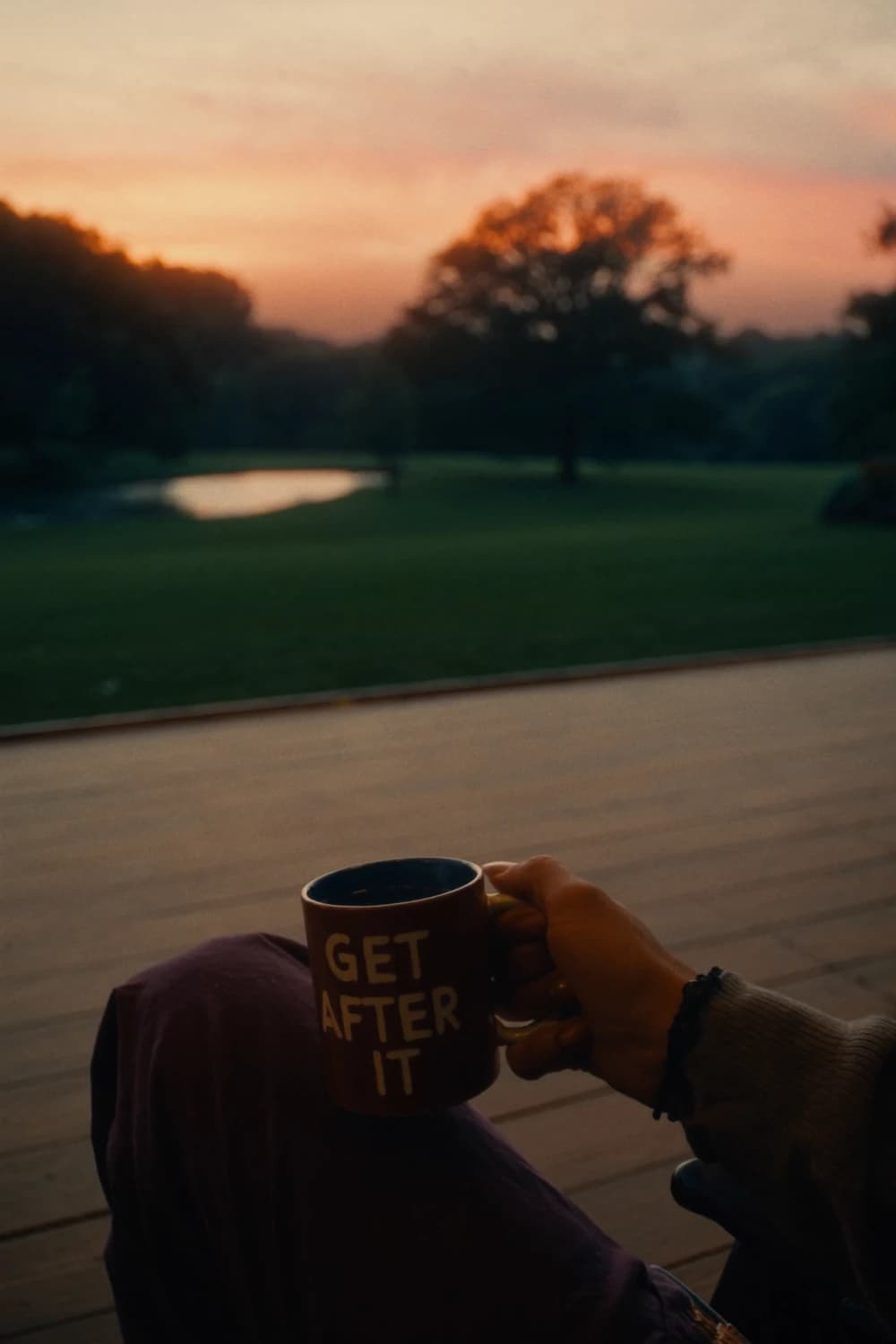 A hand holding a mug of coffee on a porch at sunrise, looking out over a park and pond