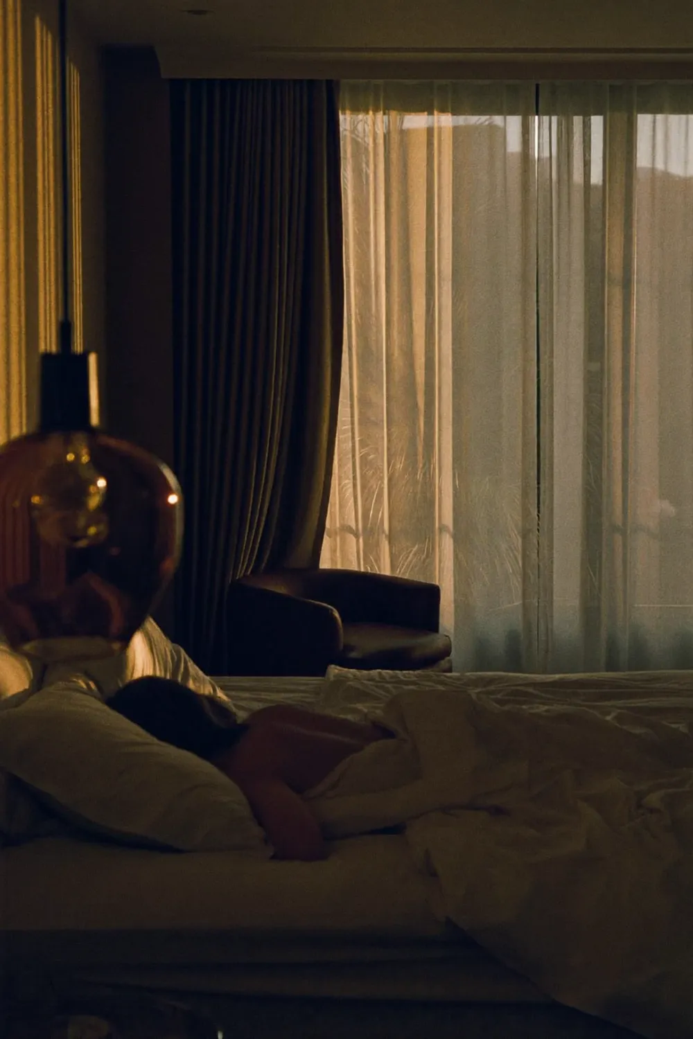 Someone asleep in a dim bedroom, soft evening light through sheer curtains