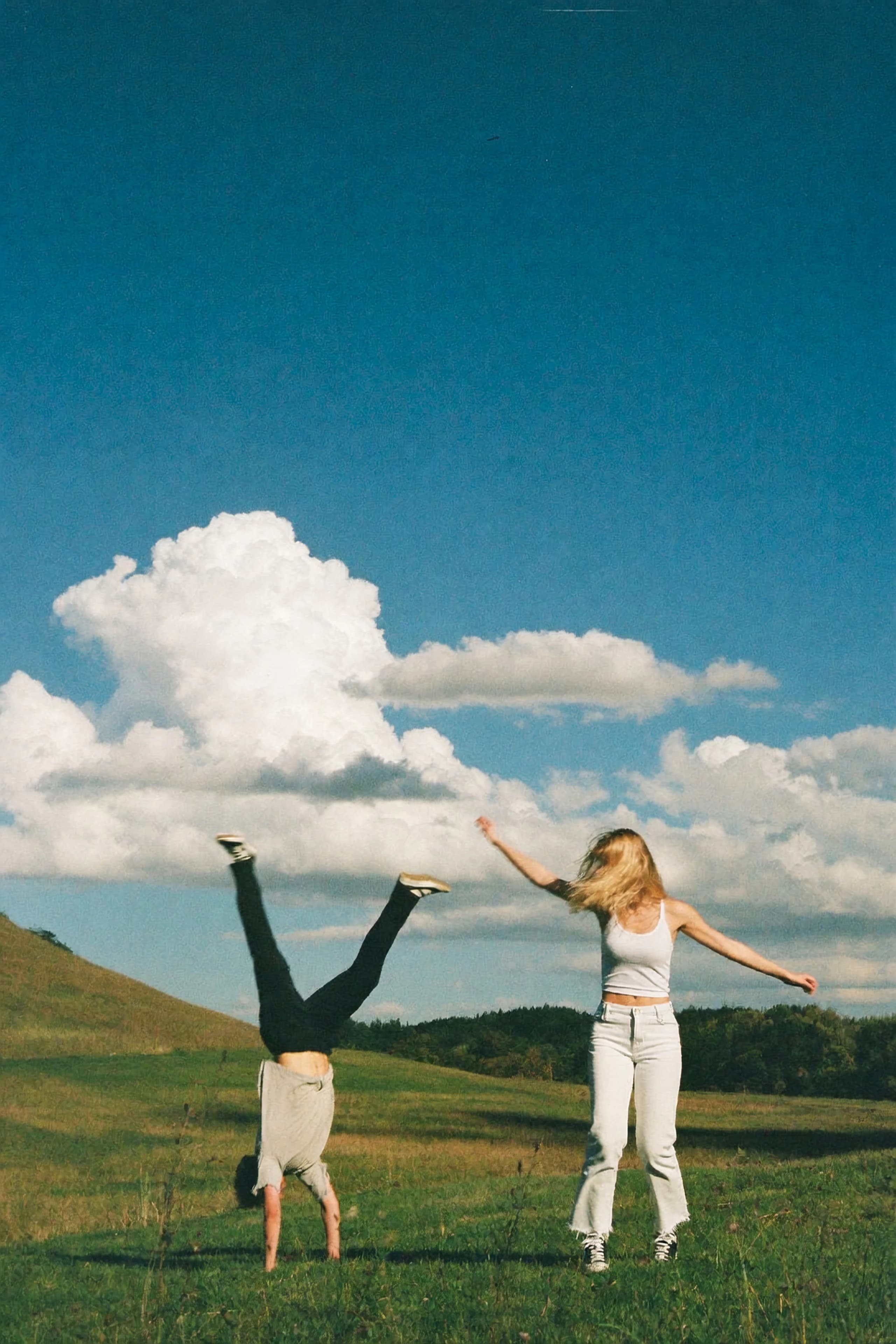 One person doing a handstand and another dancing in a green meadow under a blue sky with clouds